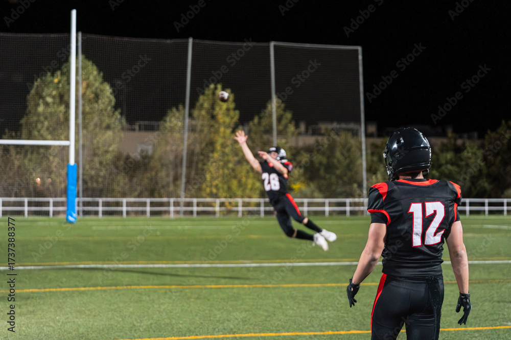 Fototapeta premium American football player in black jersey jumping for a catch at night on a green field with goalpost and teammate watching
