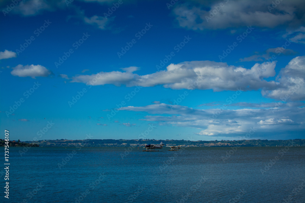 Fototapeta premium A pair of seaplanes rest on the calm waters of Lake Rotorua under dramatic storm clouds, ready for adventure against the rugged backdrop of New Zealand volcanic landscape and distant rolling hills.