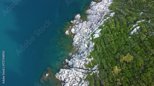 This drone footage captures the rugged, rocky coastline meeting the vibrant, clear turquoise waters of the Mediterranean Sea, surrounded by lush green vegetation. Ideal for travel, nature, and documen