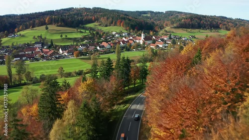 Ultra-wide aerial drone video tracks forward over a vibrant autumn european landscape with fiery red and golden trees, a winding road, cars, and a quaint village under bright daylight. Tranquil