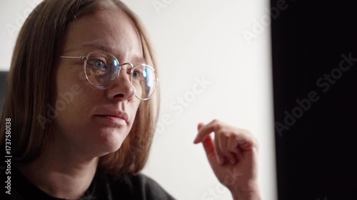 Middle aged fresh-faced caucasian woman woman with glasses, trading on Forex at nigh from home in a cozy outfit. Dimly lit home office, minimalist background. Financial chart reflexion in glasses.