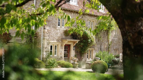 A large stone house with a large tree in front of it. The house has a lot of windows and a large front door. The tree is full of leaves and branches, and it is casting a shadow on the ground
