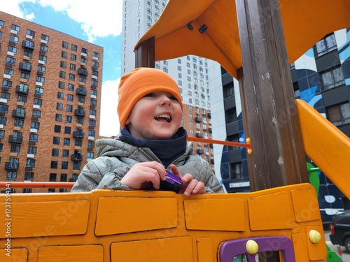Smiling young boy in orange hat enjoys outdoor play on a colorful playground structure surrounded by modern city apartment buildings. A child aged 4 years (four years old)