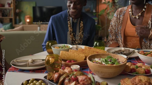 Medium shot of young Black woman placing corn on colorful Kwanzaa table as she joining family to celebrate with traditional food, unity, and cultural heritage in warm, festive home setting