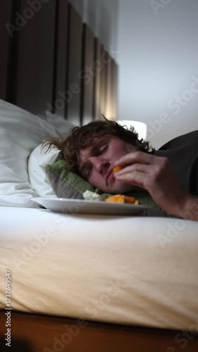 Vertical screen young man lying on bed with food plate falling asleep after eating late at night symbolizing unhealthy nutrition habits midnight snacking laziness and careless lifestyle at home