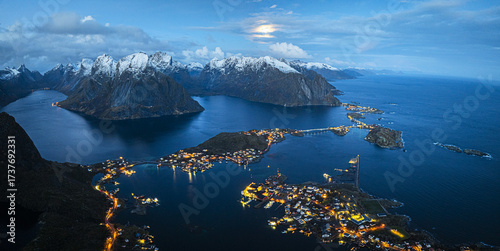Aerial view of the twinkling lights of Reine bay contrast against the dark waters and snow-capped mountains under a dusky sky, Reine, Nordland, Norway.