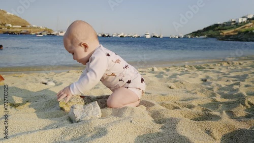 Baby exploring sandy beach, playing with small rocks, surrounded by gentle waves and distant boats, capturing the joy of childhood and connection with nature