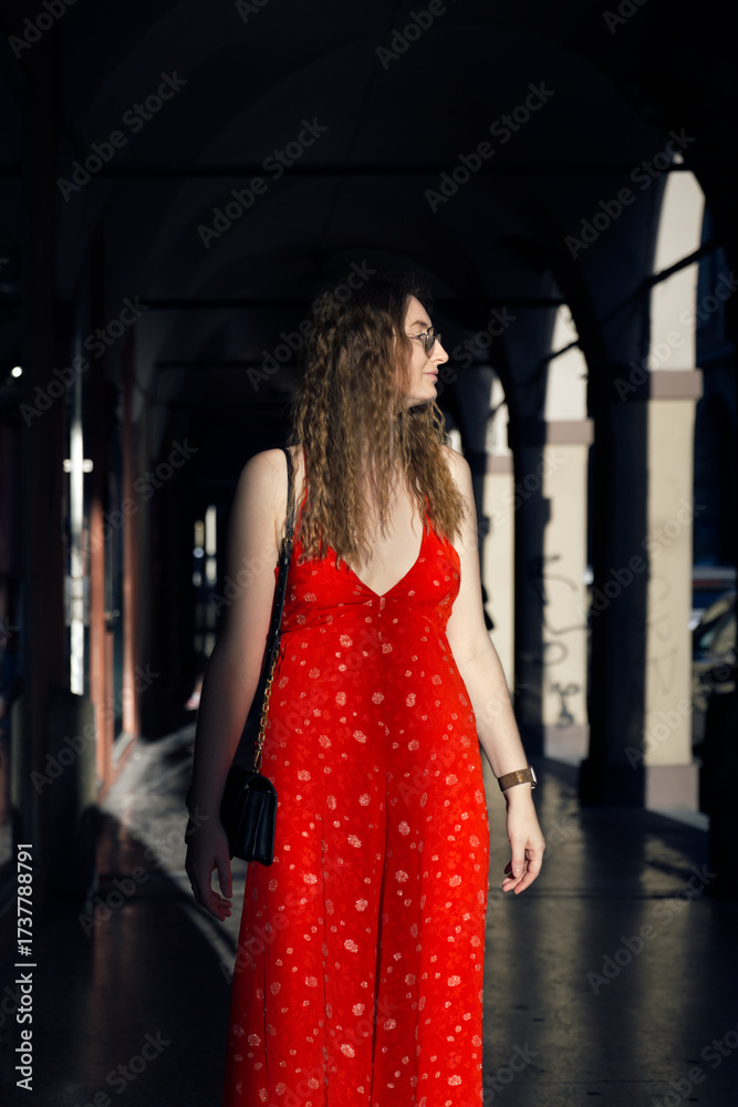 Obraz premium A woman in a vibrant red sundress with a small white floral pattern walks through a dark, arched corridor, bathed in a ray of sunlight.