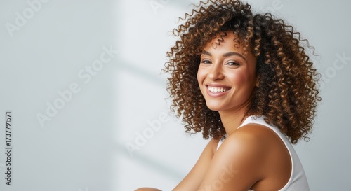 Cheerful young woman with voluminous curly hair smiling brightly at the camera. She wears a white tank top against a soft neutral background. Perfect for lifestyle, beauty, and wellness content.