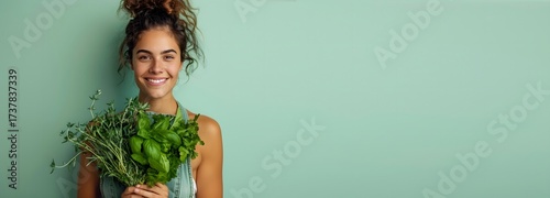 A cheerful woman showcases a collection of fresh herbs, including basil and thyme, while standing in a brightly lit indoor space. She has curly hair and radiates positivity in a casual outfit