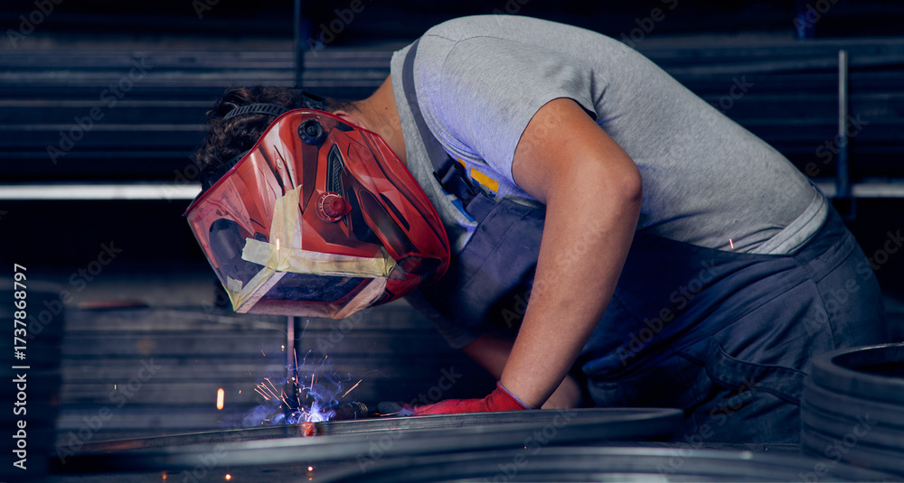 Fototapeta premium Skilled factory worker welding metal parts while wearing protective mask, gloves, and safety gear in an industrial workshop with sparks flying.