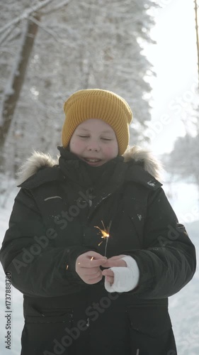 Vertical portrait of boy holding sparkler in casted arm in snowy forest. Despite his injury and adversity, happy child rejoices in approach of Christmas and believes in miracles.