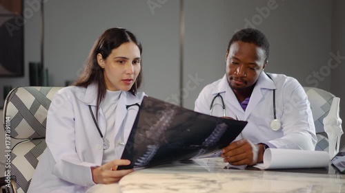 Two diverse doctors collaborate in a medical hospital, examining test results and discussing X-ray scans and use tablet computer. They focus on patient health and treatment strategies.
