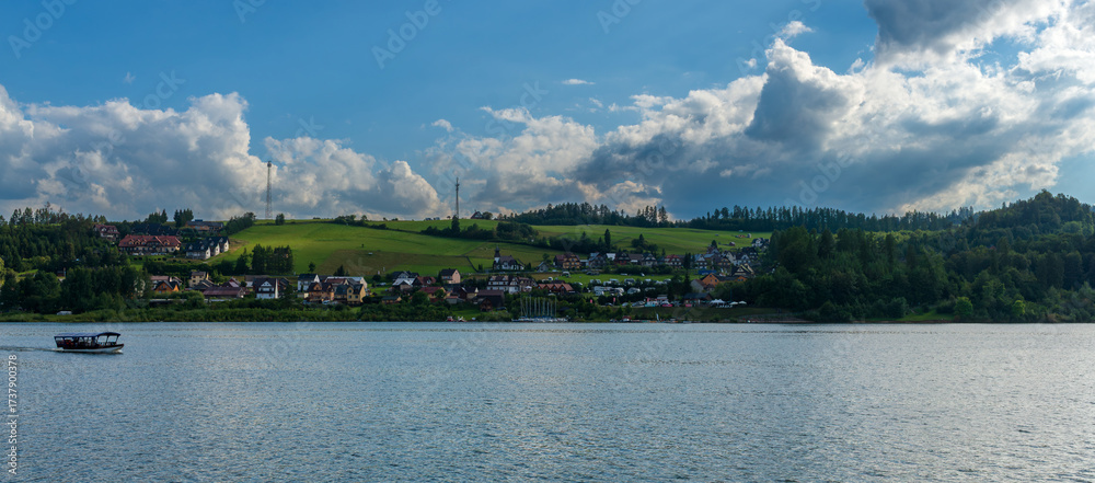 Fototapeta premium A panoramic view of a lakeside village with vibrant houses scattered across green hills. Calm water reflects the clouds and sky, creating a peaceful rural scene on a bright summer day.