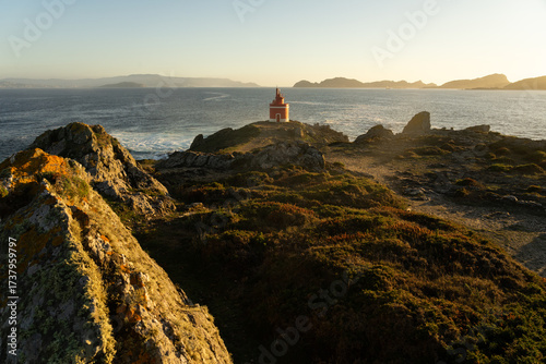 Punta Robaleira red lighthouse in Home cape at sunset in Rias Baixas zone in Galicia coast with Cíes islands in the background.