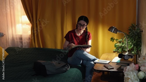 Pensive female student indoors at desk with lamp light works on academic tasks in evening time preparing for exams and university education.