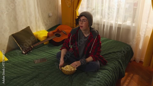 Young Caucasian woman in casual clothes and glasses sits on bed eating popcorn while watching television and checking smartphone, concept of teenage leisure at home