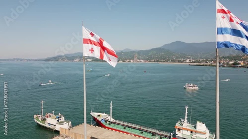 Aerial view of Batumi port with Georgian and Batumi city flags on flagpoles, ships docked in the harbor, coastline and mountains in background, Georgia