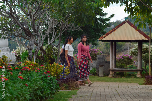 Three teenage girls in traditional skirts walk toward the upper temple garden, chatting together near a gazebo and decorative plants while appreciating the natural surroundings.