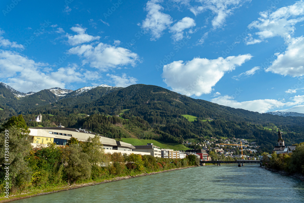 Fototapeta premium Scenic view of alpine town with river, church tower, and mountains in the Austrian Alps.