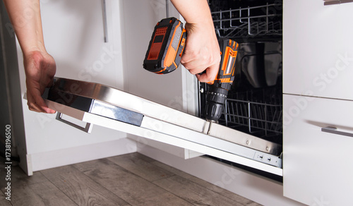 The hands of a craftsman with a screwdriver install a kitchen front panel on a dishwasher, industry