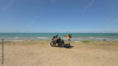 Motorcycle parked by the sea on a sunny day, facing the blue horizon.
