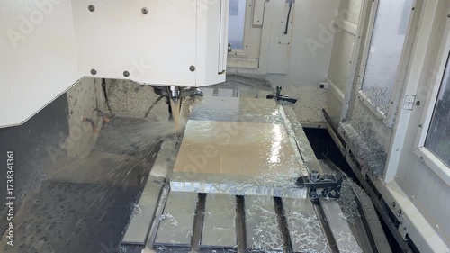 Wide view of CNC milling under flood coolant: a face mill cuts an aluminum plate as streams of emulsion and chips splash across the T‑slot table and fixtures inside the machine enclosure.