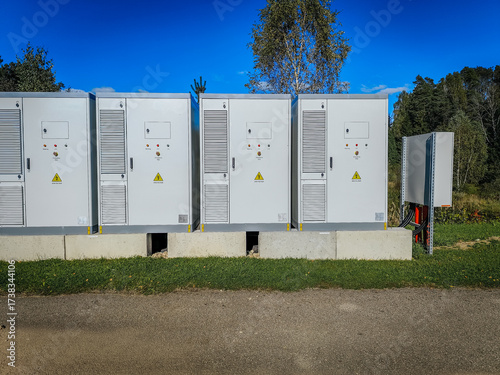 A row of outdoor high-voltage electrical cabinets with warning labels, ventilation panels, and emergency stop buttons under a clear blue sky.