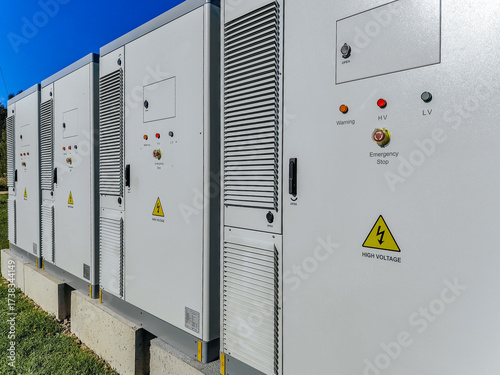 A row of outdoor high-voltage electrical cabinets with warning labels, ventilation panels, and emergency stop buttons under a clear blue sky.