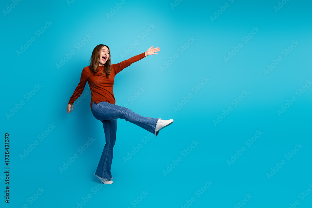 Obraz premium Joyful young woman wearing a brown sweater and jeans, posing with a kick motion against a blue background