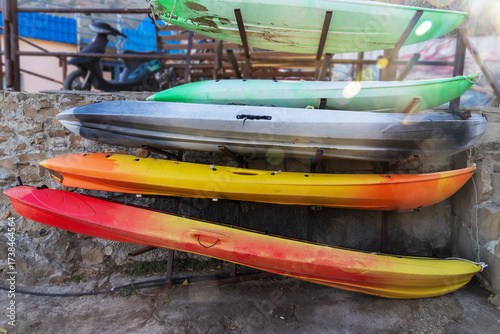 Colorful kayaks stacked on stone wall, vibrant red, yellow, orange, green, and gray boats stored outdoors in bright sunlight.