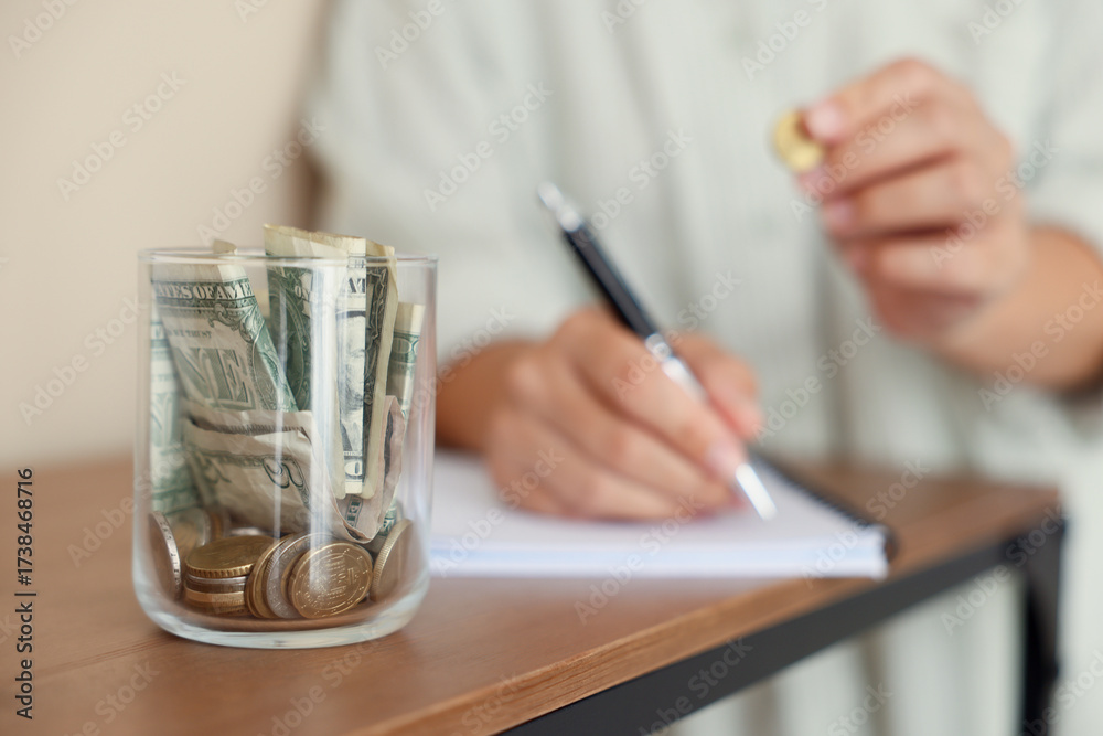 Fototapeta premium Woman putting money into jar and writing in notebook at wooden table indoors, selective focus