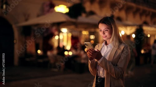 Woman standing on a city street at night, looking at smartphone with warm smile, illuminated by screen light