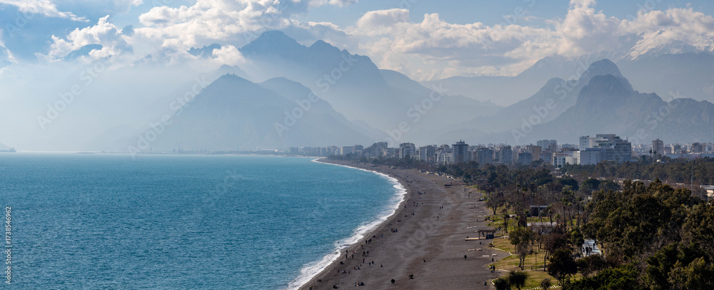 Fototapeta premium Panoramic view of Konyaaltı Beach's curved coastline with turquoise sea, white foam, and people relaxing against a backdrop of hazy Taurus Mountains. Sunny winter day in Antalya, Turkey.
