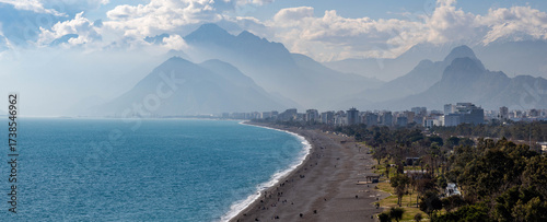 Panoramic view of Konyaaltı Beach's curved coastline with turquoise sea, white foam, and people relaxing against a backdrop of hazy Taurus Mountains. Sunny winter day in Antalya, Turkey.

