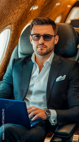 A smiling businessman in glasses and a brown jacket works on a blue laptop in the cabin of a private jet. The atmosphere is one of success, comfort, and productive travel.