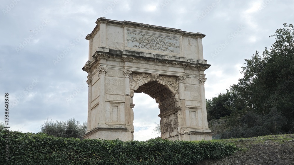 Fototapeta premium Rome, Italy - 13 January 2025. The Arch of Titus stands alone on a grassy rise, with its single barrel-vaulted passage, inscription, and carved reliefs still intact against a cloudy sky.