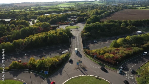High-angle aerial shot capturing a busy motorway intersection with an overpass and a large, circular roundabout, surrounded by green and autumn-colored trees and fields.