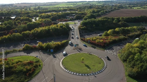 A high-angle aerial view of a busy motorway junction featuring a large roundabout and an overpass, surrounded by dense trees with touches of autumn color and rolling fields in the distance.
