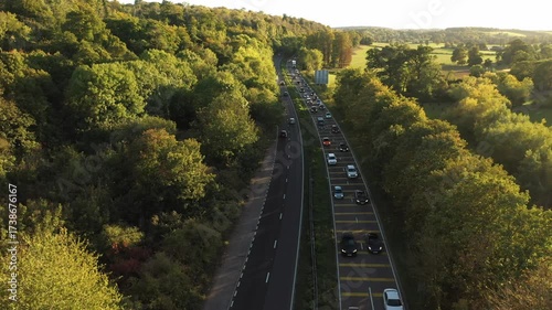 A panning aerial shot revealing a long stretch of dual carriageway with traffic backed up, situated between a dense, bright forest on one side and open, sunlit parkland on the other, under a bright sk