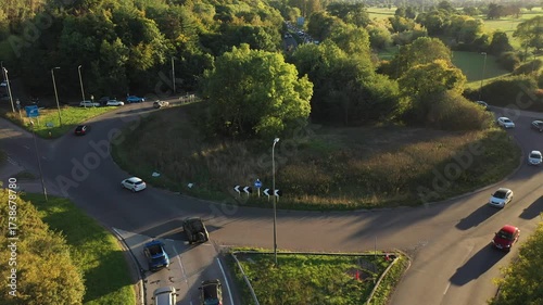 An aerial shot descending onto a large roundabout at a busy junction, showing cars navigating the circle, surrounded by thick green trees with the main road extending into the distance.