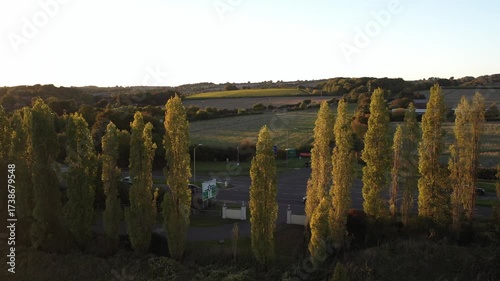 An aerial tracking shot that moves backward, pulling away from a row of tall, slender trees, revealing a road entrance, a small parking area, and the open, rolling countryside bathed in the golden lig