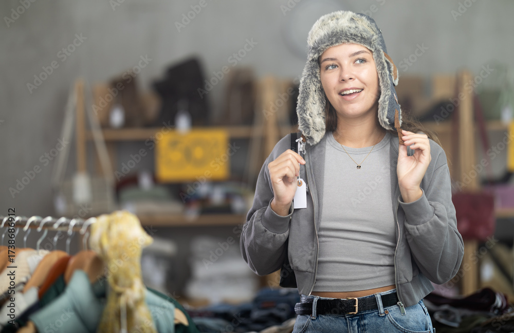 Fototapeta premium Positive young girl trying on fur hat while shopping in clothing store