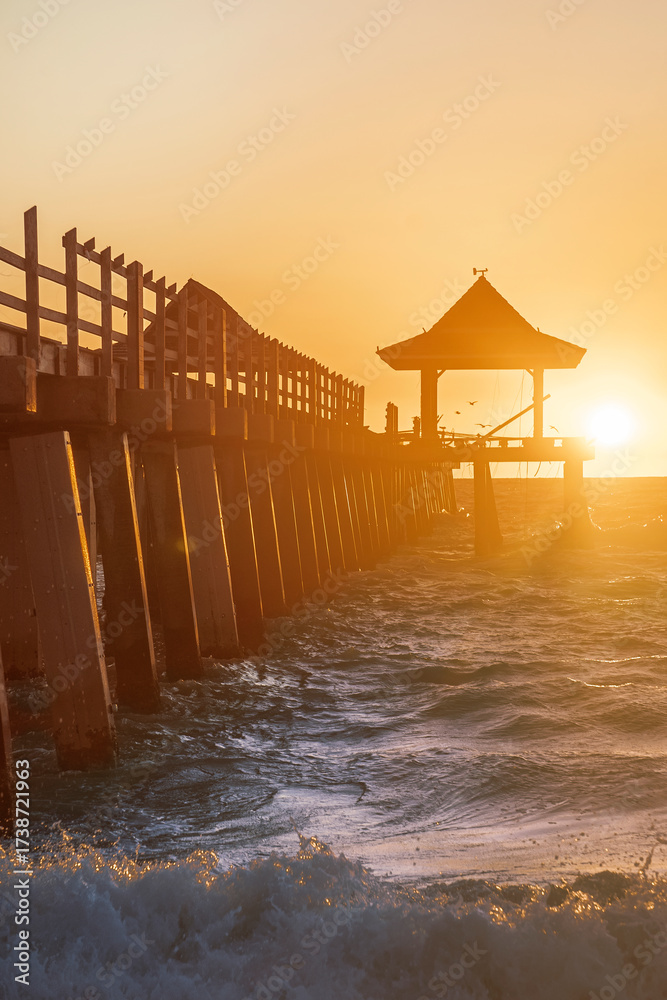 Fototapeta premium Waves crash against the Naples Beach Pier at Golden Hour Sunset, Florida, USA