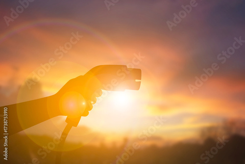 Hand Holding Electric Vehicle Charging Plug Against Sunset Sky