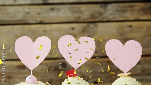 Gold confetti drifting over wooden wall, pink hearts on sticks rising, presenting cupcakes on tray