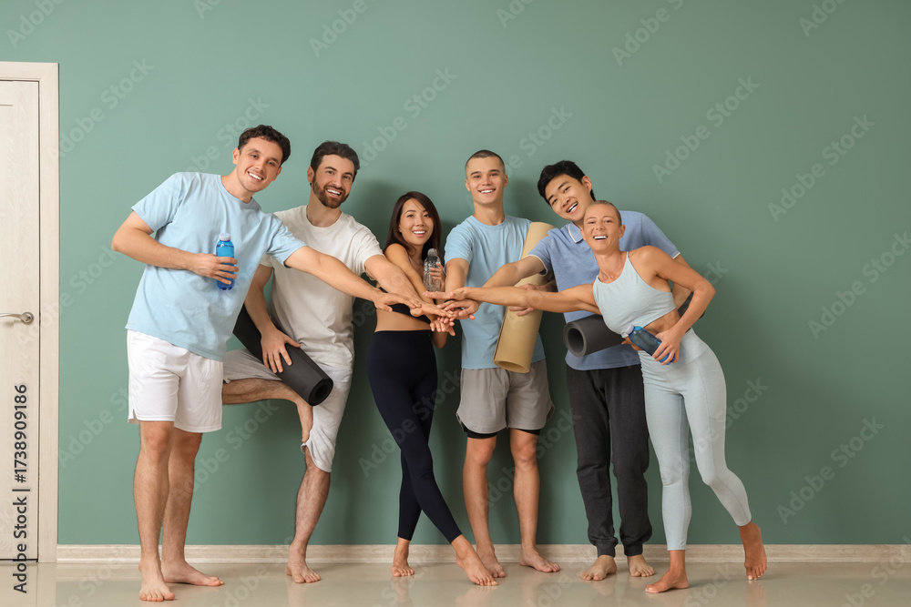 Fototapeta premium Group of sporty people with yoga mats and water bottles putting hands together near green wall