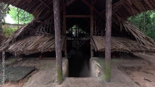 Entrance to an underground room at the Cu Chi Tunnels in Vietnam, with a traditional thatched roof shelter and narrow earthen pathway.