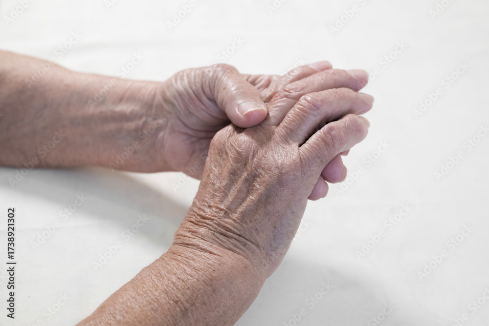 Fototapeta premium Two hands of an elderly person. The skin of the hands is thin, with pronounced wrinkles and veins, which indicates age. One hand holds the other. The color scheme of the photo is neutral, with light s