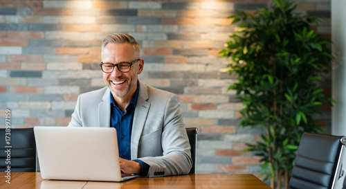 Happy mature businessman working on laptop in office, wearing glasses and smiling, with brick wall background